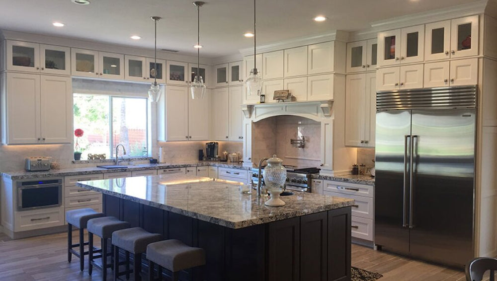 Upscale kitchen with white custom cabinets, oversized granite island with seating, pendant lights, built-in range hood, and stainless steel French-door refrigerator.