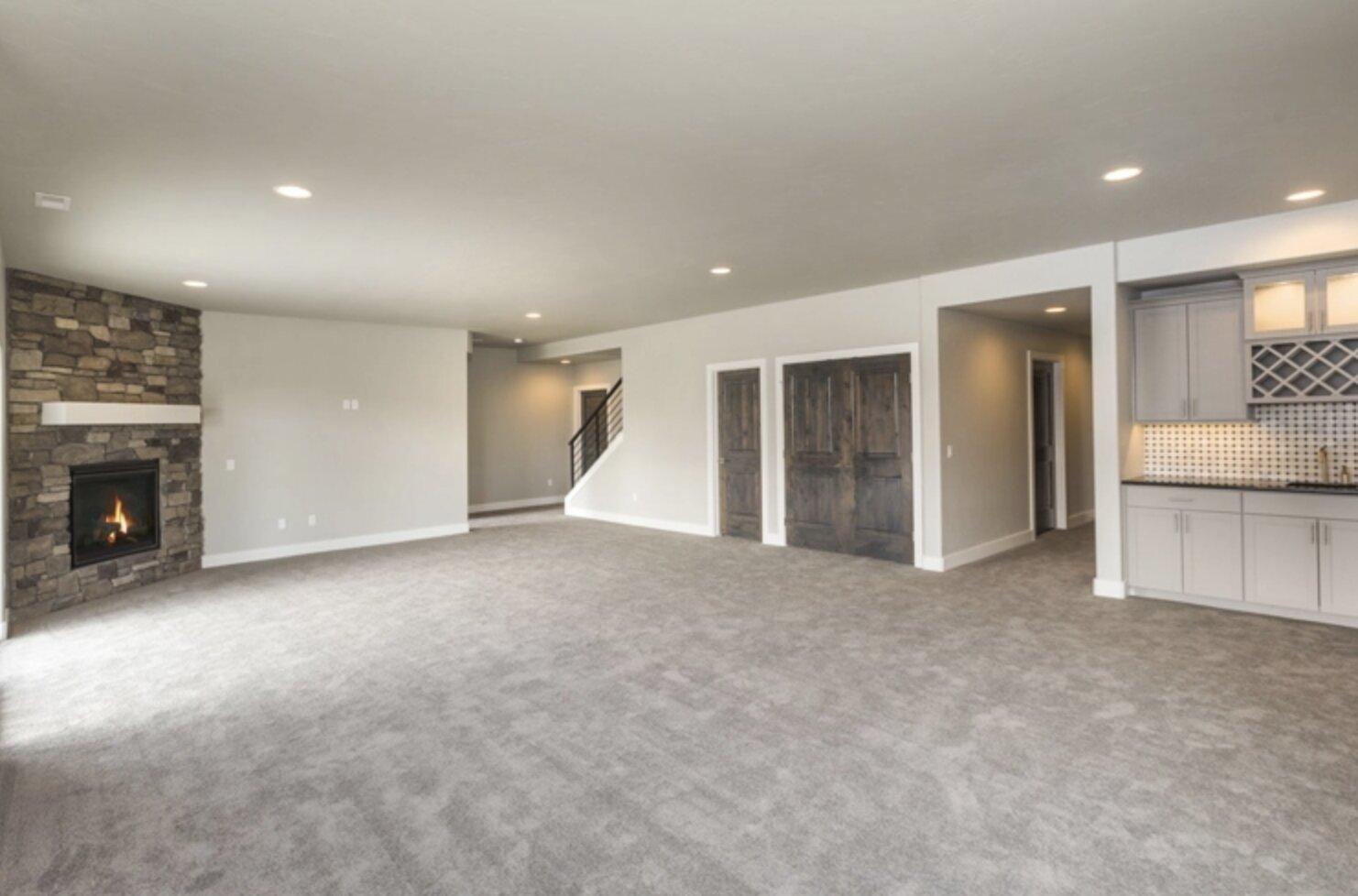 Large finished basement with gray carpet, recessed lighting, stone fireplace with gas insert, rustic double barn doors, and a built-in wet bar with cabinets and backsplash.